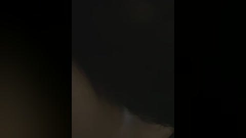 A dimly lit video featuring a dark, shadowy figure in the center, possibly a person or object, with indistinct features due to low visibility. The background is predominantly dark, with hints of light and shadow.