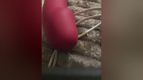 Video of a red vibrator with white wires on a beige, textured fabric surface. The image is cropped and focused closely on the device, emphasizing its smooth, glossy texture.