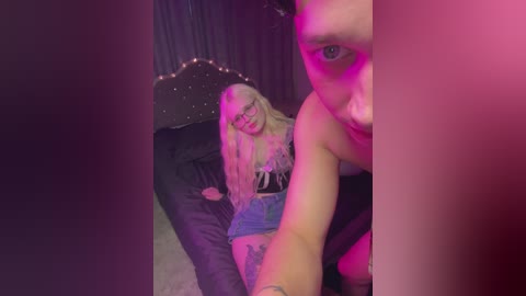 A video of a blonde woman with glasses, wearing a black corset, sitting on a black beanbag chair, surrounded by pink and purple lighting, with a tattoo on her thigh.