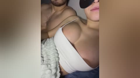 Media: A video of a topless woman with medium-sized breasts wearing a white crop top and a beige knitted blanket. A man with a beard is partially visible, and both have fair skin.