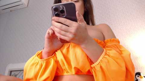 Media: Video of a woman in a bright orange off-shoulder top, holding a smartphone, with a white textured wall and air conditioning unit visible in the background.