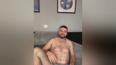Media: A video of a bearded, nude, fair-skinned man with a muscular build, seated on a gray couch, smiling. Background features framed art and a white light switch.