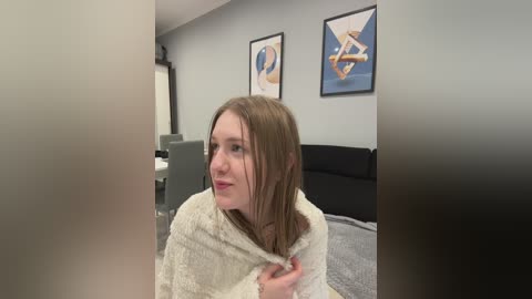 Media: Video of a young woman with fair skin and shoulder-length brown hair, wrapped in a white towel, standing in a modern living room with grey walls and black furniture, featuring abstract artwork.
