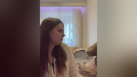 Video of a young woman with long brown hair, wearing a white robe, gazing out a window with sheer curtains. Bedroom background features a bed with rumpled sheets and pillows.