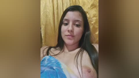 Video of a young woman with long dark hair, fair skin, and small breasts, topless, holding a blue and white towel. She sits against a golden-yellow textured backdrop.