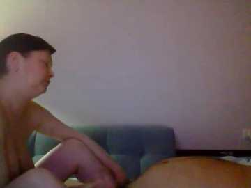 A dimly-lit video of a young man with short dark hair, shirtless, sitting on a blue couch with a white brick wall background.