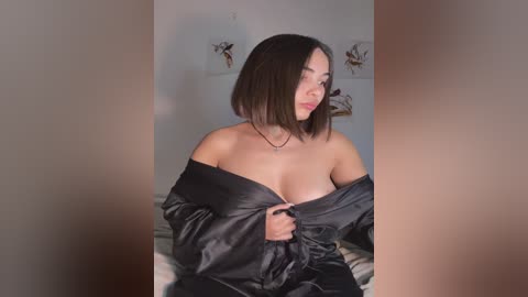 Media: Video of a young Asian woman with straight, shoulder-length black hair, wearing an off-shoulder black satin robe, partially revealing her medium-sized breasts, in a dimly lit bedroom with minimalist decor.