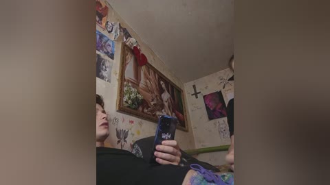 A video of a person with short hair, holding a phone, in a cluttered room with colorful posters and graffiti on the walls, wearing a black shirt.