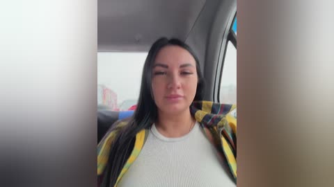 Video of a young woman with long black hair, light skin, and a slight smile, wearing a white ribbed top and a yellow plaid shirt, seated in a car.
