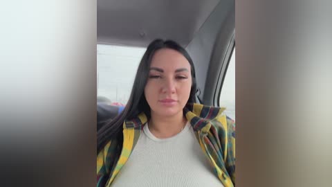 Video of a young woman with long black hair, fair skin, and a neutral expression, wearing a white ribbed top under a plaid shirt, seated in a car.