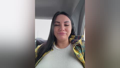 Video of a woman with long black hair, wearing a yellow and green plaid shirt over a white ribbed tank top, smiling with eyes closed, inside a car.