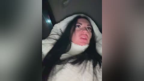 A video of a woman with long, dark hair, wearing a white turtleneck, covering her face with both hands, appearing distressed, in a dimly lit room.