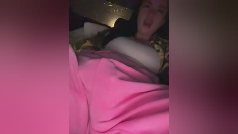 A video of a woman lying on a bed, partially covered by a pink blanket, her mouth slightly open, with a blurred, dimly lit background.
