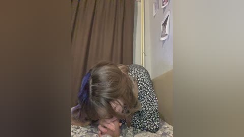 Media: Video of a woman with short brown hair, wearing a floral dress, lying face down on a bed, covered with a patterned blanket, in a dimly lit room with beige walls and brown curtains.