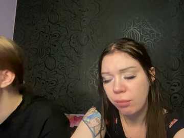 A video of a young woman with light skin and long brown hair, wearing a black shirt with colorful patches, sitting indoors with a dark, patterned wall behind her.