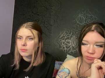 Media: Video of two young women with fair skin and long hair, one with a nose ring, sitting on a bed with dark floral wallpaper.