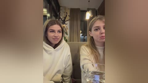 Media: Video of two young women with blonde hair, wearing casual outfits, sitting at a dining table in a dimly-lit, cozy restaurant. One makes a kissy face, while the other smiles.