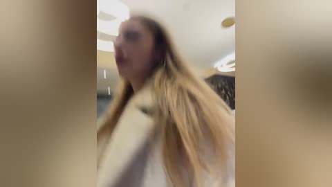 Media: A blurred video shows a blonde woman with long hair, possibly in a white coat, walking indoors with a slight smile. The background includes a ceiling and light fixture.
