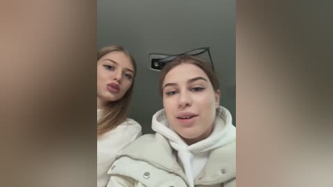Media: Video of two young women, one with long blonde hair, the other with brown hair, both wearing white hoodies, posing indoors with blurred background.