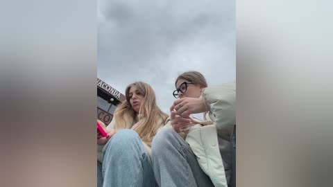 Media: Video of two young women, one with long blonde hair and glasses, the other with brown hair, sitting on a bench in a cloudy outdoor setting.