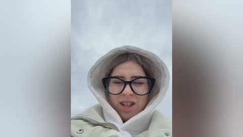 Media: Video of a young woman with light skin, wearing black-rimmed glasses, a white hoodie, and a beige jacket, standing in a snow-filled, blurred background.