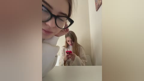 Media: Video of a blonde woman with glasses, wearing a white sweater, looking at a red phone in a mirror, reflecting a beige wall with framed art.