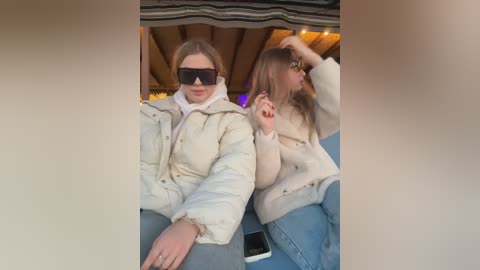 Media: Video of two women, one with light brown hair, sunglasses, and beige puffer jacket, the other with blonde hair, wearing a beige sweater, seated in a cozy outdoor setting with wooden beams.