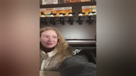 Media: A video shows a smiling woman with curly blonde hair, wearing a beige sweater and a gray coat, standing in front of a coffee machine with \"Buddy\" coffee bags.