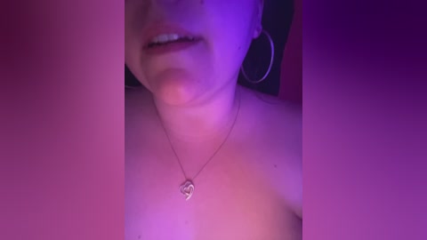 A dimly lit, close-up video of a fair-skinned woman with a silver heart pendant necklace, wearing hoop earrings. The image is framed by a pink and purple gradient background, focusing on her partially visible face and shoulders.
