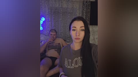 Video of a young woman with long black hair and fair skin wearing a gray shirt with \"Aloha\" printed on it, seated in a dimly lit room with a man in the background.