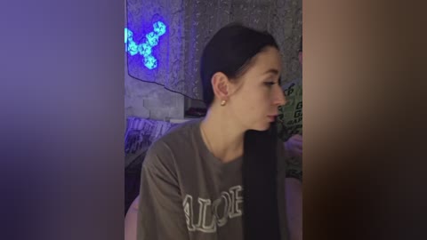 A video of a young woman with fair skin and long black hair, wearing a gray \"LOVE\" t-shirt, sitting indoors with a blue-lit background and a textured wall.
