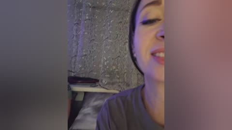 A video of a young woman with light skin and dark hair, smiling slightly, taken from a slightly awkward angle. She wears a grey T-shirt. The background features a textured, grey wall and a white shelf with a black object.