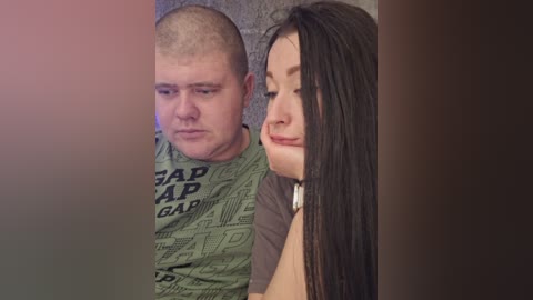 A video shows a man with a shaved head wearing a green GAP t-shirt and a woman with long dark hair in a brown shirt, both with neutral expressions, in a dimly lit room.