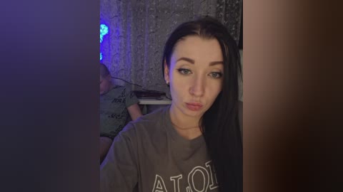 Video of a young woman with long black hair, wearing a grey t-shirt, indoors with dim lighting, blurred background, making a pouty face.