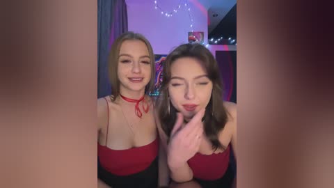 Media: Video of two young women with fair skin, wearing red tops, one with a red choker, smiling, indoors with a festive background.