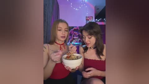 Media: Video of two young women, one blonde in a red dress, the other brunette in a red dress, eating ramen in a dimly lit room with colorful posters and string lights.