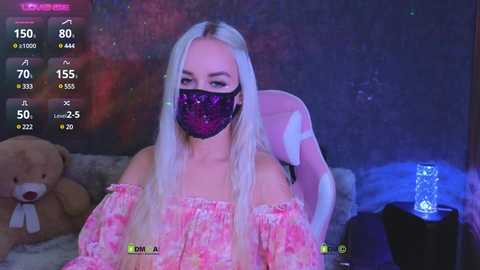 Video of a young woman with long platinum blonde hair, wearing a pink floral off-shoulder dress, black face mask, and sitting in a plush pink chair. Background includes a teddy bear and blue glowing lamp.