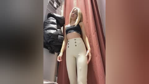 Media: Video of a slim, blonde woman in a black crop top and high-waisted white pants standing in a dressing room with a puffy jacket hanging and a red curtain.