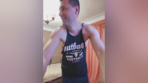 Media: Video of a smiling man in a black \"Uttah\" tank top, standing in a beige-walled bedroom with a bed and orange curtains.