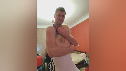 Media: Video of a shirtless man with a lean physique and fair skin, covering his chest with his right arm while wearing only white boxer shorts. Background shows a messy bedroom with orange walls and scattered items.