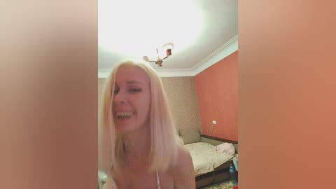Media: Video of a blonde woman with fair skin, wearing a pink top, laughing in a dimly-lit bedroom with an unmade bed and orange walls.
