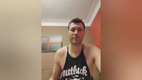Media: Video of a young, light-skinned man with short brown hair, wearing a black tank top with \"outdoor\" text, standing in a room with beige wallpaper, a framed painting of a lighthouse, and orange wall.