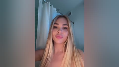 Media: Video of a young woman with long, straight blonde hair, fair skin, and light makeup, standing in a bathroom with white shower curtain. She has a neutral expression and is partially obscured by the frame.