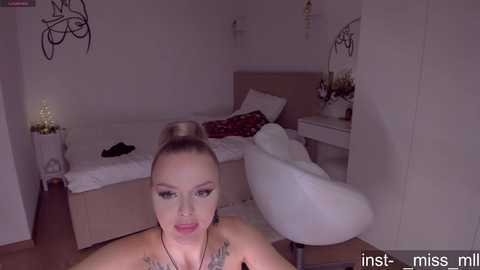 Video of a woman with blonde hair in a bun, wearing makeup and a black top, in a minimalist bedroom with a white bed and wall art.