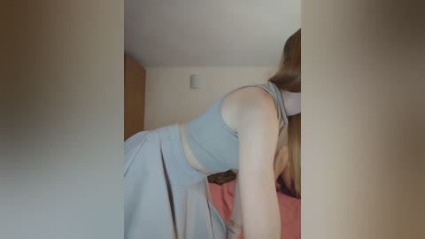Video of a young woman with long, straight light brown hair, wearing a light blue tank top and matching skirt, bending over a bed with pink sheets in a dimly lit room.