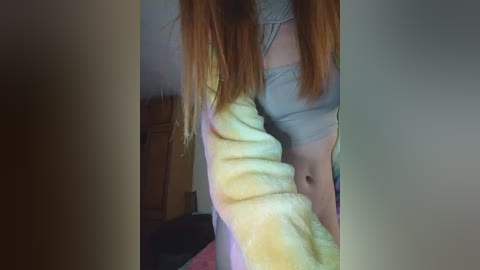 Video of a young woman with fair skin and long, straight red hair. She's wearing a light grey crop top and a fluffy, yellow bathrobe, with a hint of a wooden dresser in the background.