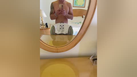 Video of a shirtless man with a tattooed arm, wearing white boxer shorts, taking a mirror selfie in a hotel room with a wooden desk, lamp, and framed artwork in the background.