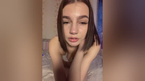 A video of a nude, fair-skinned, young woman with straight, dark brown hair, large brown eyes, and a slender build, kneeling on a bed, taken from a close-up, slightly blurred perspective.