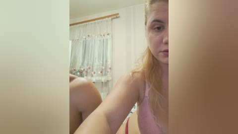 A video of a fair-skinned, blonde woman in a pink bra, sitting in a bedroom with floral curtains and a window.