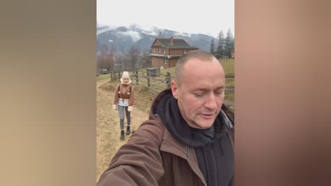Media: Video of a smiling bald man in a brown jacket and black scarf walking on a dirt path, with a blonde woman in a brown jacket and white hat behind him, in a mountainous, snowy landscape.